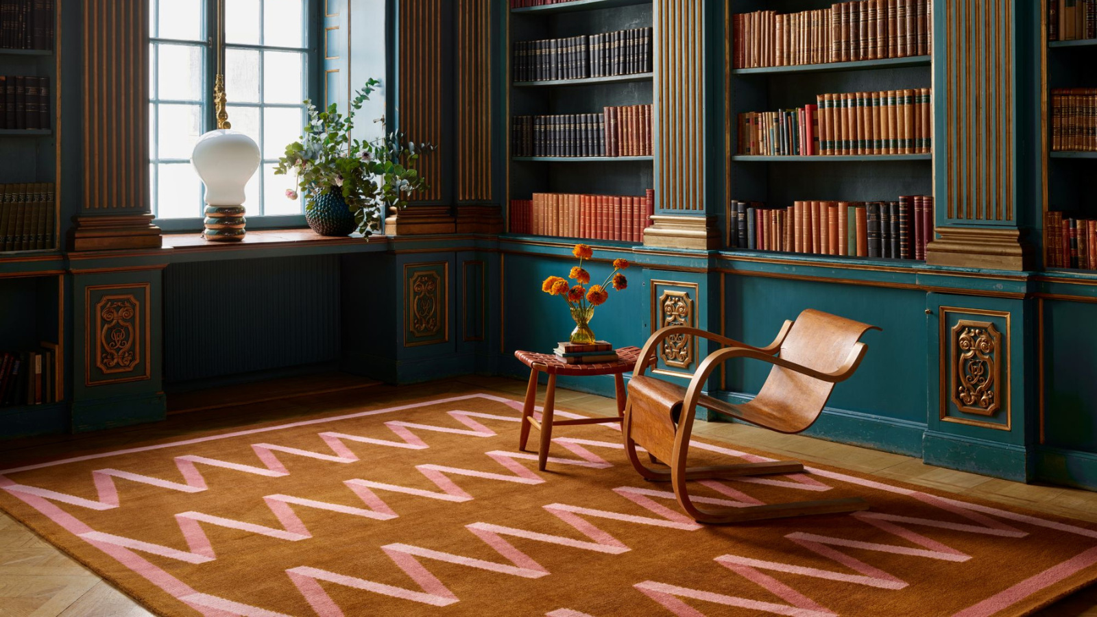 a study room with a large rug with pink zig zag stripes and a brown background, teal painted bookshelves holding many books, and a wooden chair and an accent table in the middle of the room
