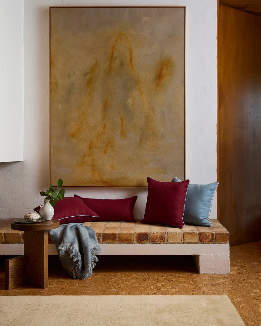 Image of a white entryway with a love white and brick bench seat that has maroon and light blue cushions on it. There is a large beige abstract art hanging on the wall.