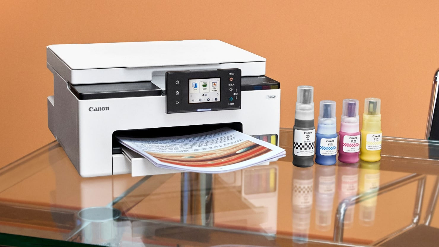 A Canon MegaTank printer with bottles of ink sitting on a glass desktop with orange background.