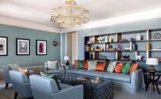 Interior view of Assila Hotel, Jeddah, Saudi Arabia featuring grey flooring, a grey rug, blue walls, blue sofas and chairs with cushions in different colours, a blue padded coffee table with items on top, framed wall art, a shelving unit filled with books, vases and other items and a gold chandelier