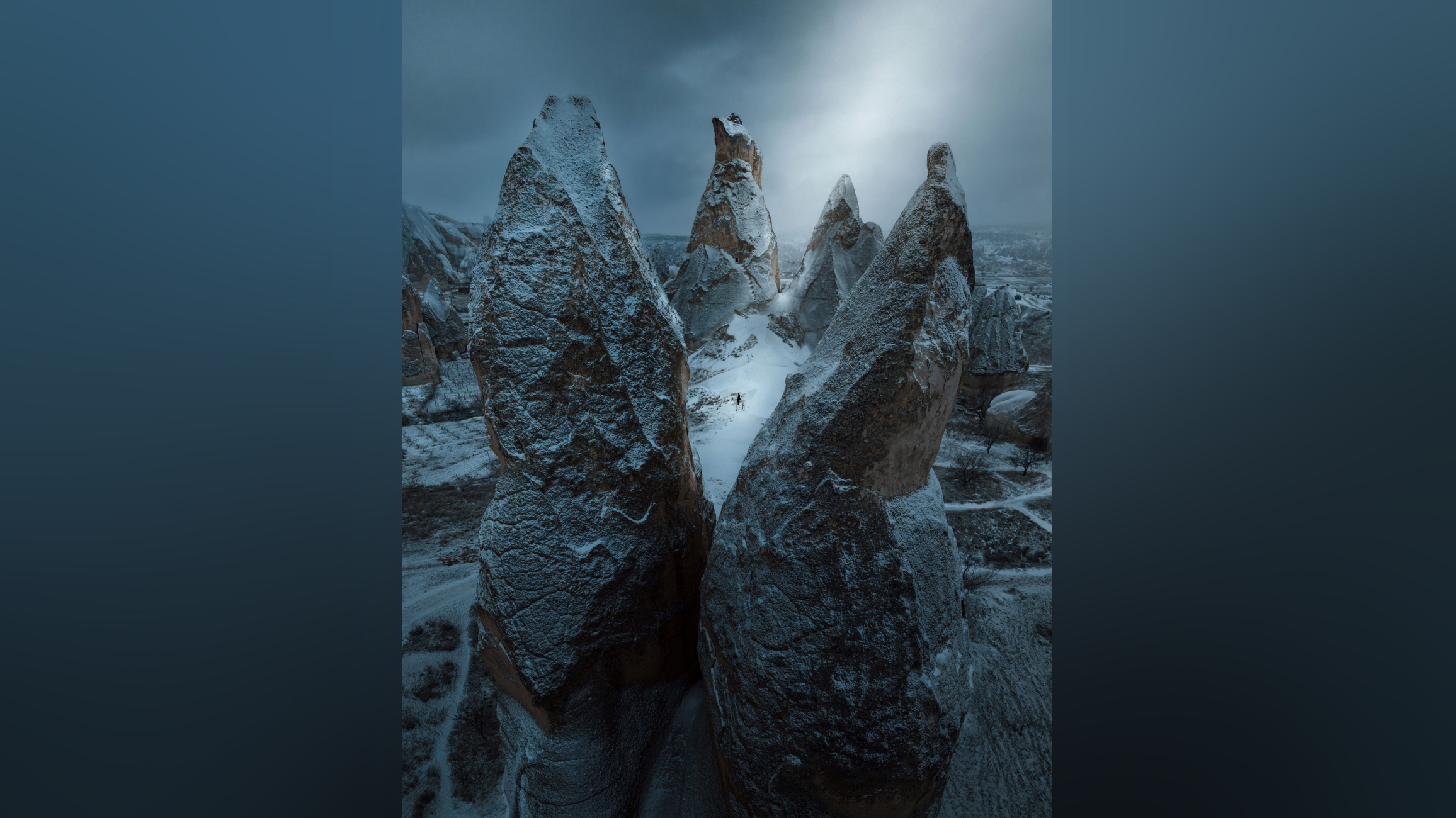 A dramatic winter landscape featuring towering rock formations covered in snow, with a lone figure standing amidst them