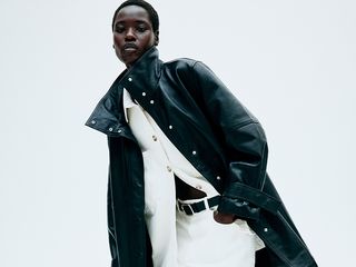 Model wears white button down, white jeans, and black jacket while bending over slightly and looking at the camera.