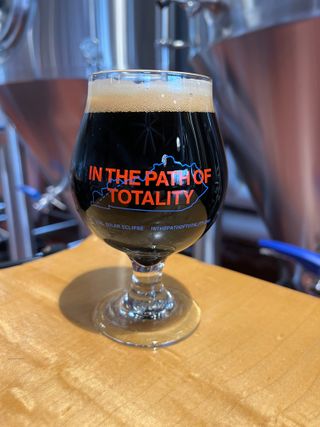 a dark beer rests in short glass, standing on a wooden table with large brewing equipment in the background.