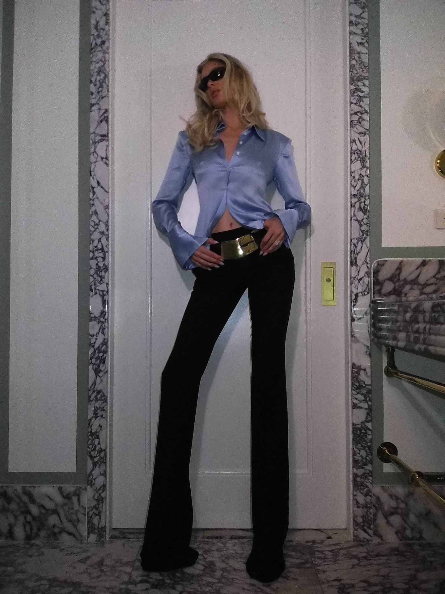 Elsa wears a blue silk button-down top, belt, and long black pants.