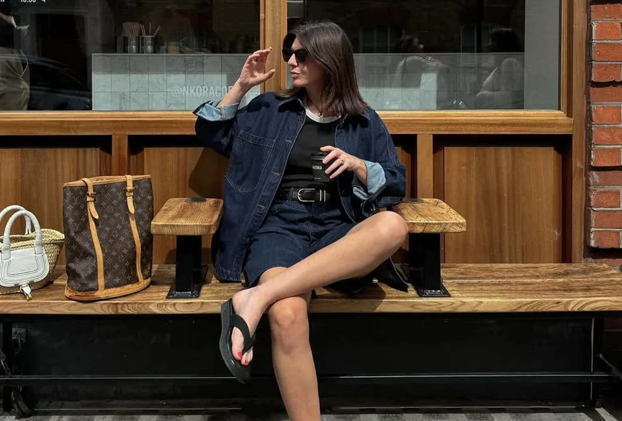 Woman wearing spring flip-flop outfit with denim jacket, denim shorts, black sweater, and black flip flops.