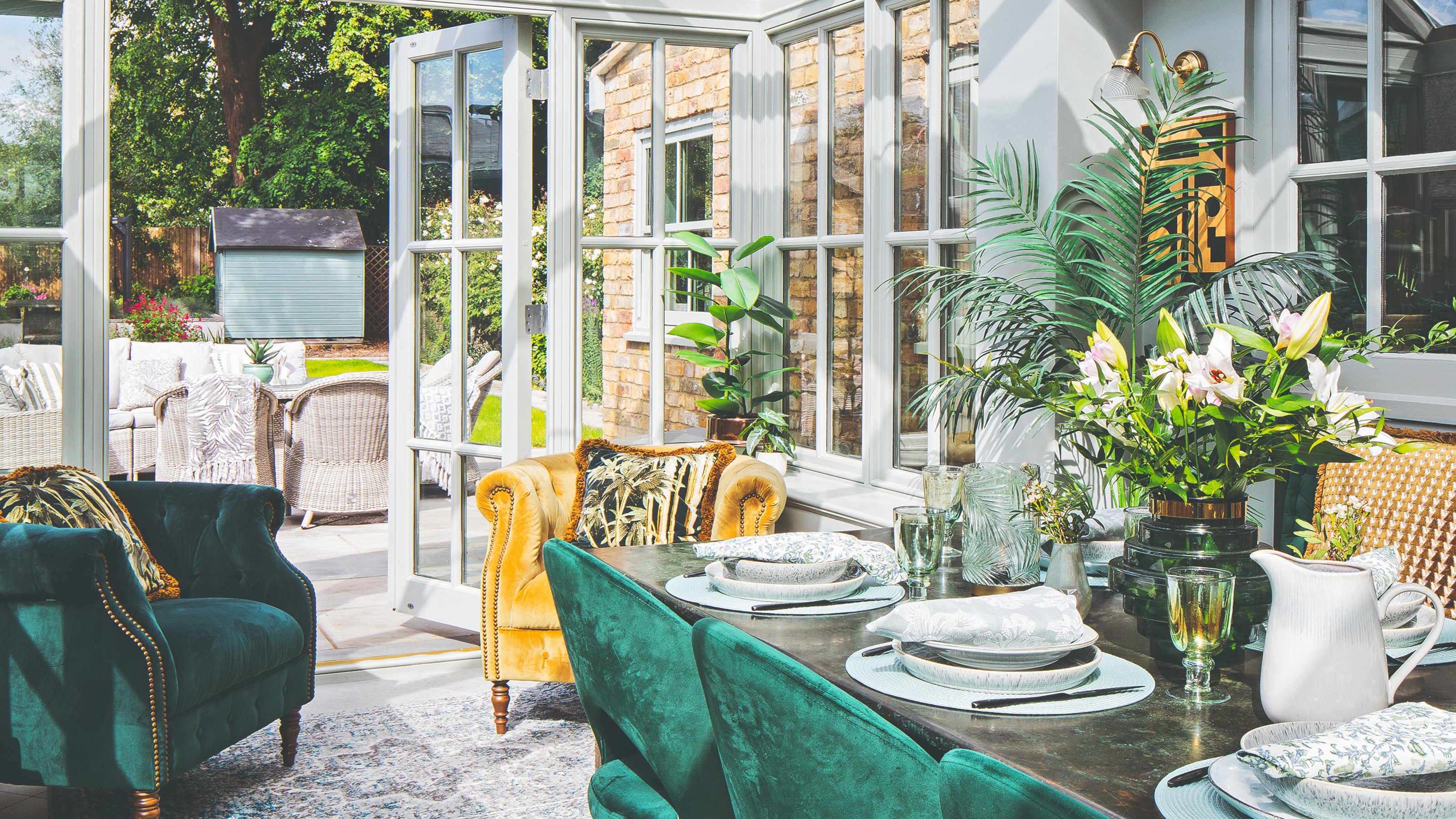 A conservatory with a dining area with emerald green dining chairs and two velvet armchairs in yellow and green