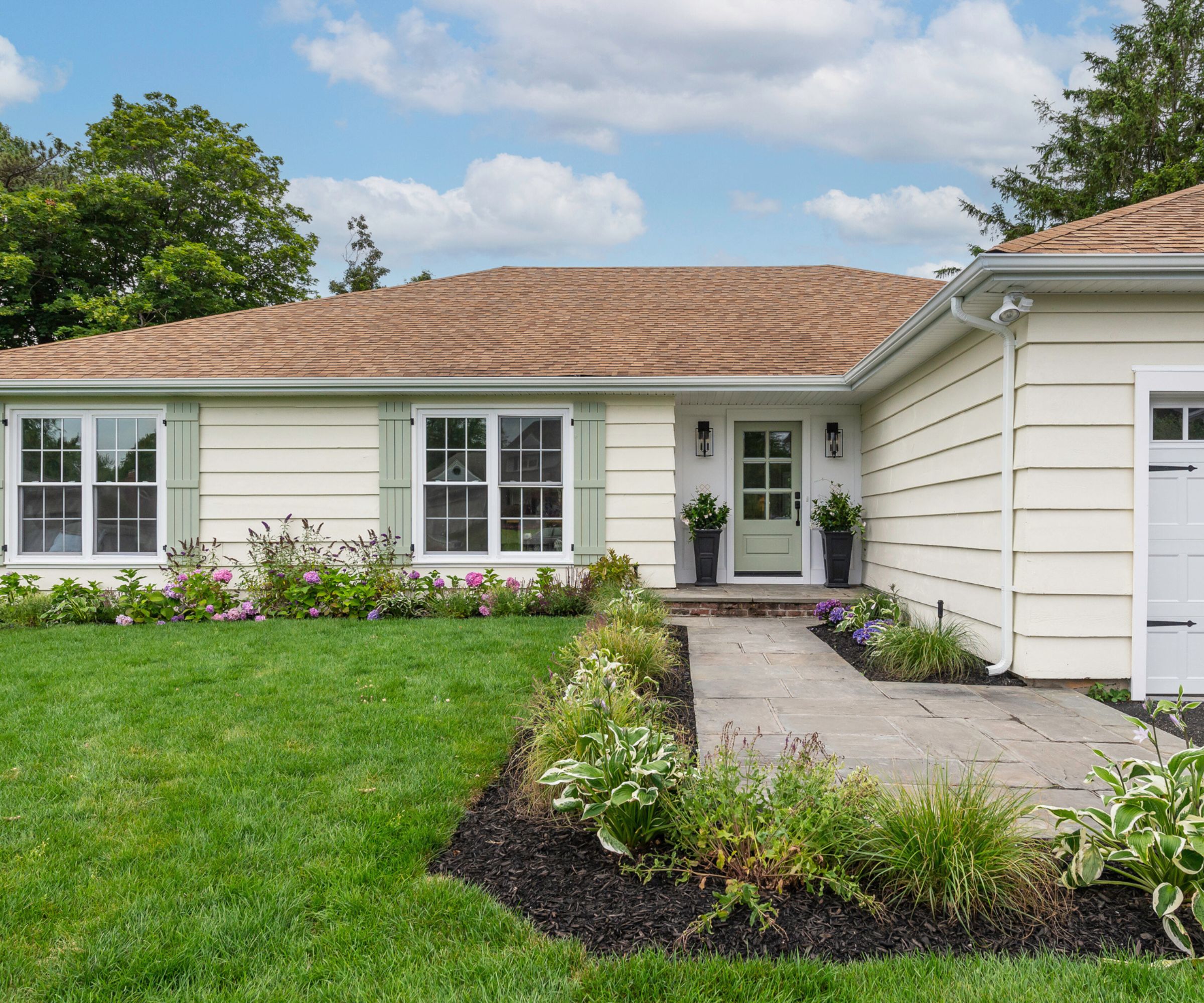 A bungalow with a creamy white exterior, light green window shutters and front door, and a red brick roof and grass lawn with plants.