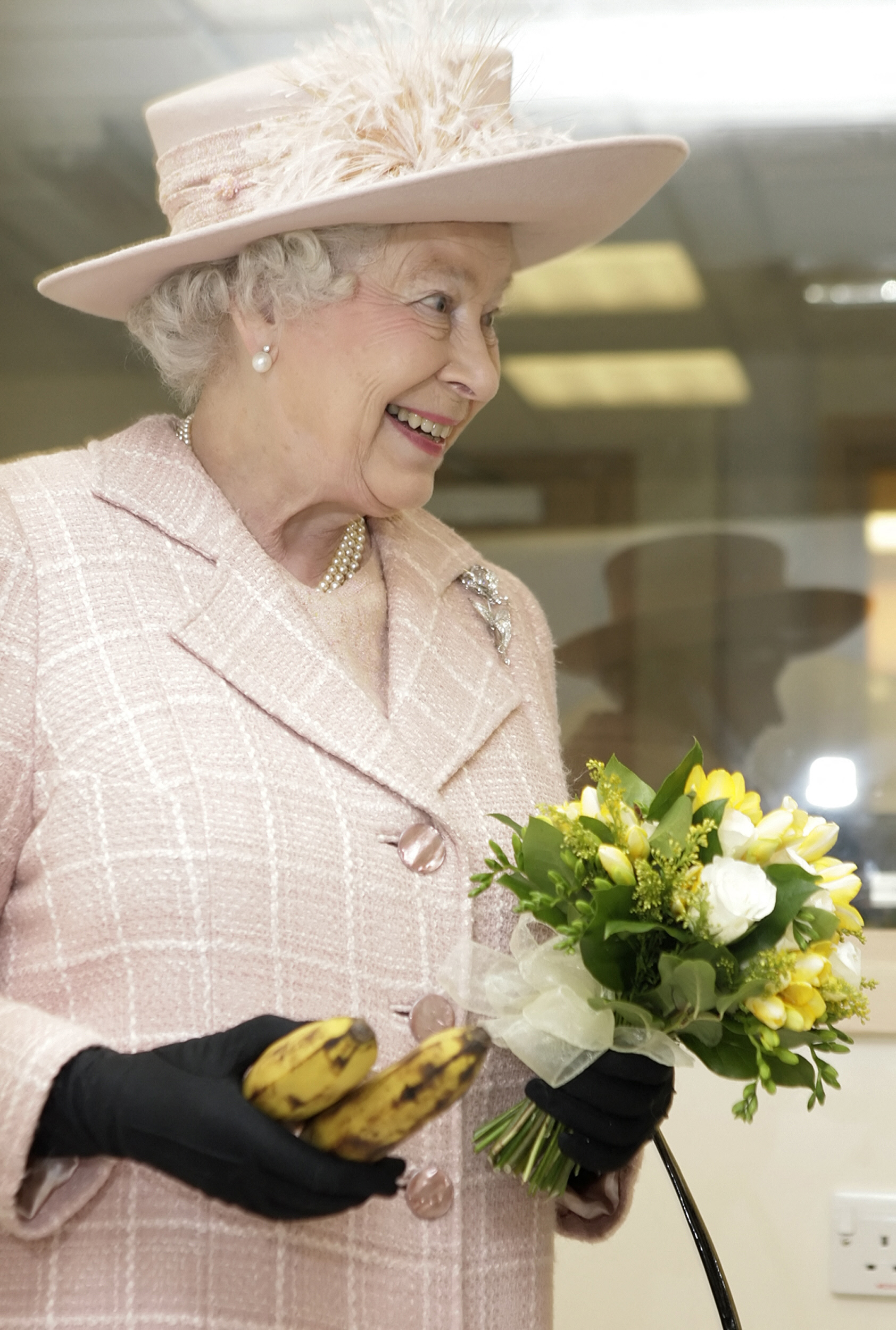 Queen Elizabeth wearing a pink suit and hat holding bananas and flowers