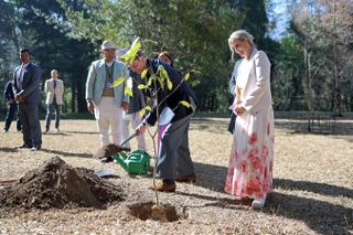 Duchess Sophie wearing a long white skirt with red flowers and a cream blazer standing behind Prince Edward,who is shoveling dirt while planting a tree in Nepal