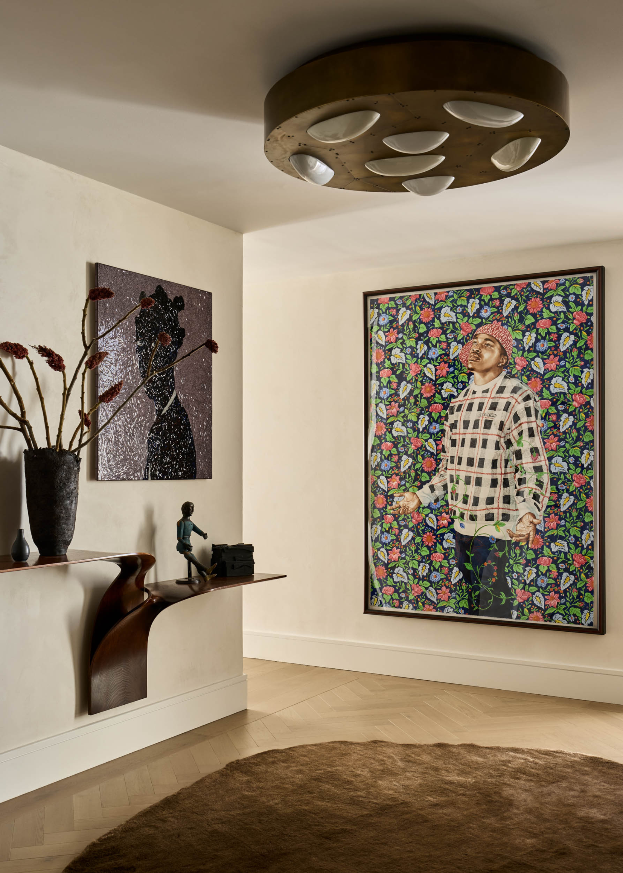 A hallway with hanging art, an overhead light feature, a round brown rug, a wall shelf with a vase and a statue