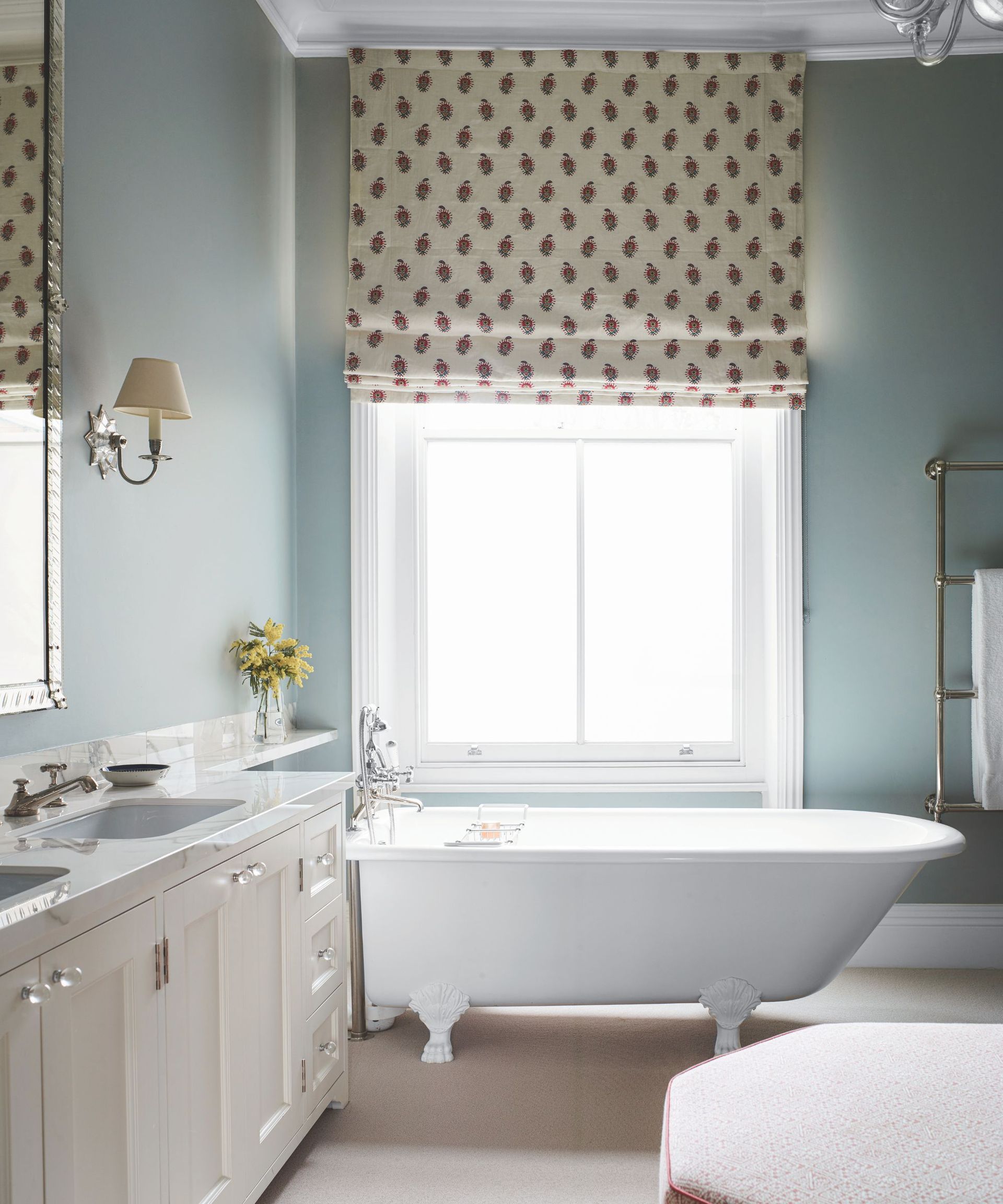 A blue bathroom with a freestanding bathtub, what vanity, and a patterned blind over the window