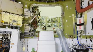 a square-shaped opening in a spacecraft surrounded by hoses, computer bits and wires. through the window is a factory facility