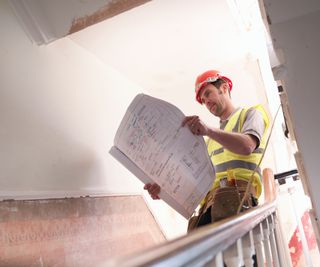 builder on stairs with yellow hi vis and red hard hat looking at set of plans