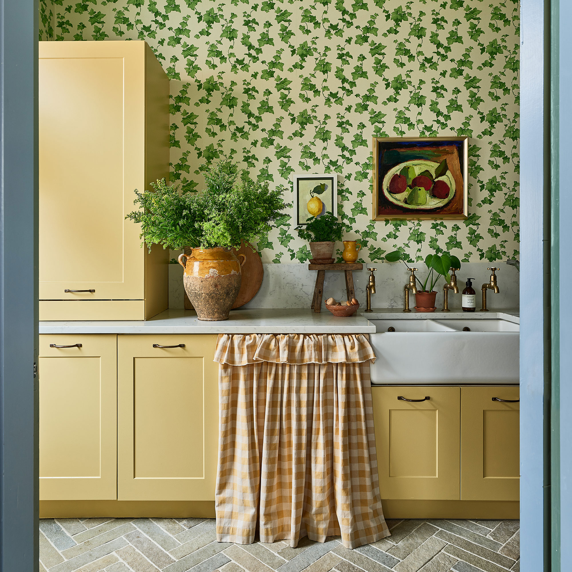 Kitchen with yellow cabinetry, ivy patterned wallpaper, stone chevron flooring and yellow gingham curtain covering appliance