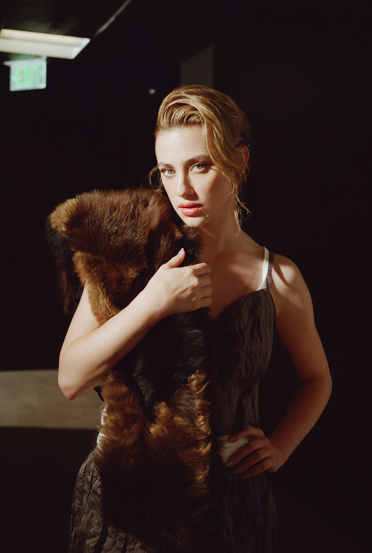 Lili Reinhart staring at the camera wearing a Miu Miu top and and skirt set and holding a fur scarf