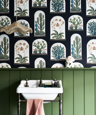 A master bathroom with lower green-painted wood paneling and a botanical wallpaper behind a traditional white sink
