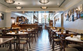 A restaurant in the Redchurch Townhouse hotel. The area is covered with round wooden tables and chairs. We can see a wooden bar far away, with rounded wooden shelves behind it.