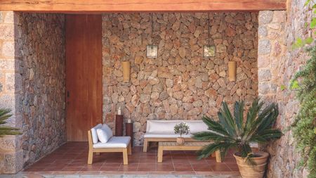 Rustic patio with a white and wooden outdoor sofa and chair, a low wooden table, a potted palm, brick walls, glass lantern lights hanging from the ceiling, and a terracotta tiled floor.