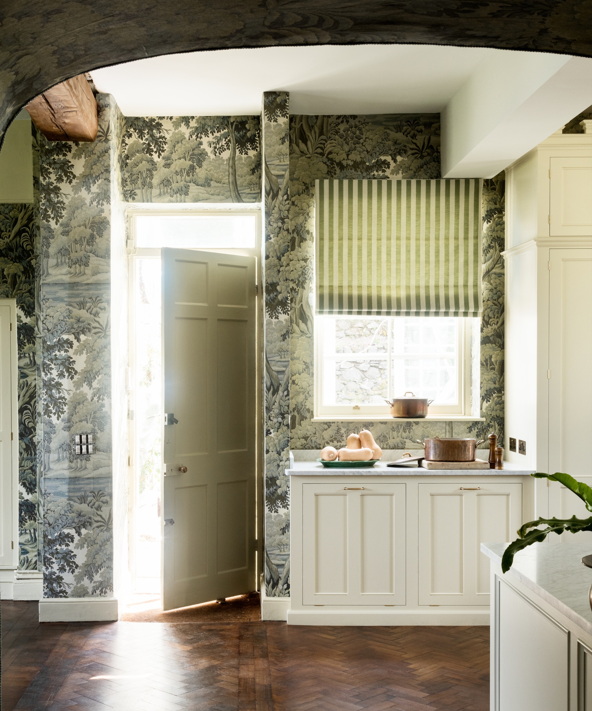 A white kitchen wallpapered in green woodland imagery, complete with striped curtains