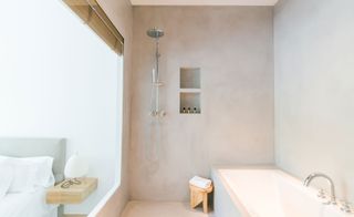 A hotel bedroom joined with a bathroom with a bed, a shower, a tub, a curved tap, a wooden bench and built in wall storage.