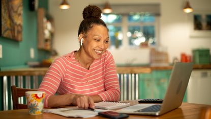 A woman smiles as she looks at her laptop while sitting at her kitchen table.