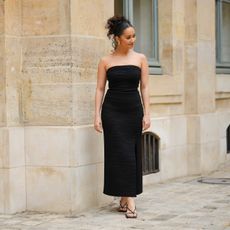 Lena Mahfouf wears gold large earrings, a black embossed striped pattern / shoulder-off / long slit / split dress, black shiny strappy leather small heels sandals , outside the COS show, on April 26, 2023 in Paris, France.