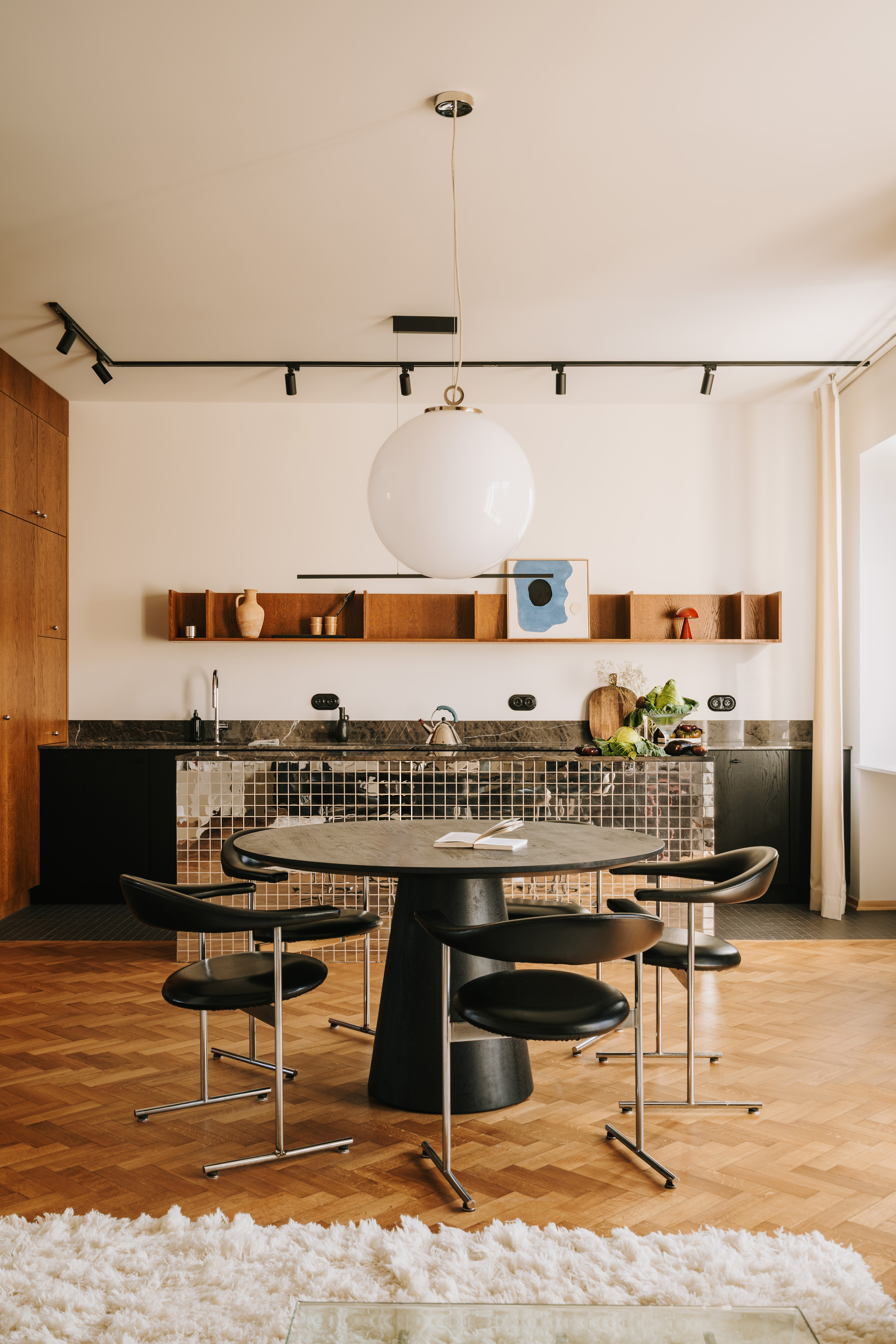 Image of a white room with wooden floors with black kitchen cabinetry and a dico ball mirrored kitchen island. There is a round black dining table with black chairs.