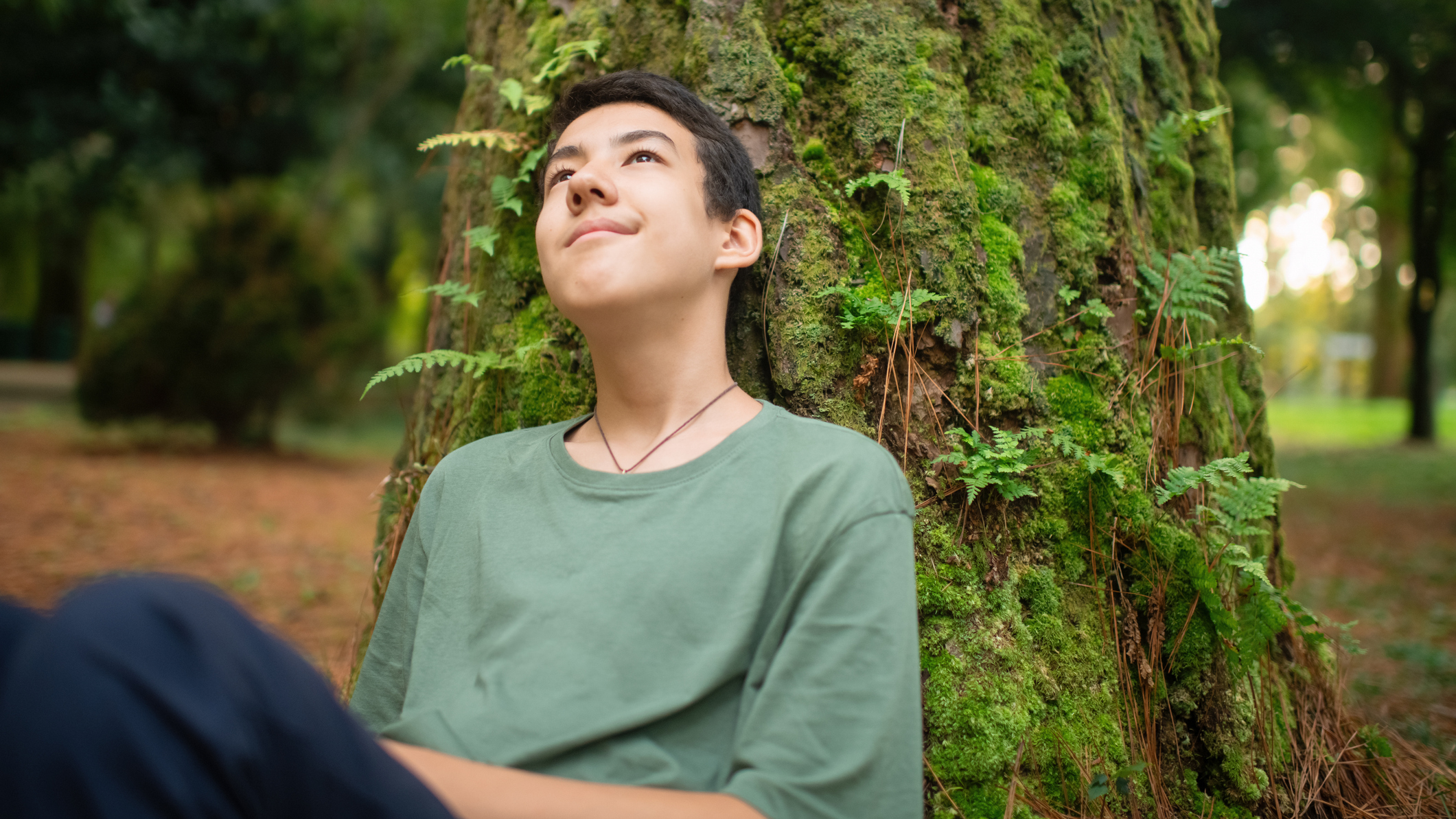 Woman sits with her back against a moss-covered tree trunk in a forest setting, looking up with an inscrutable, half-smiling expression