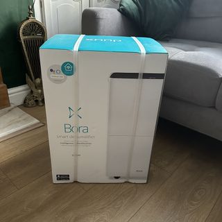 The Duux Bora Smart Dehumidifier in its packaging box in a living room with a wooden floor