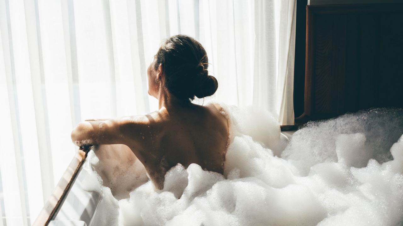 Back view of a woman sitting in a bubble bath facing a window - best bath salts