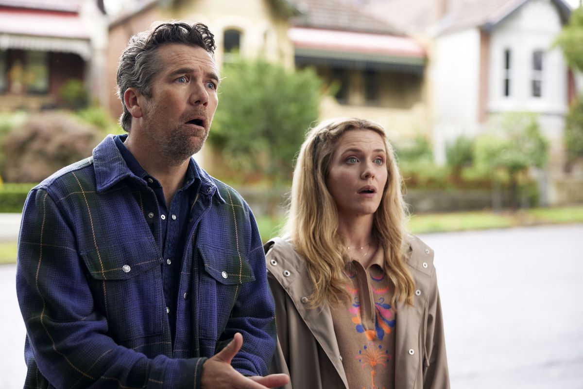 Patrick Brammall and Harriet Dyer as Gordon Crapp and Ashley Mulden in Colin From Accounts season two. They are standing on the street, talking to someone out of shot, and Gordon is reacting to them in distress