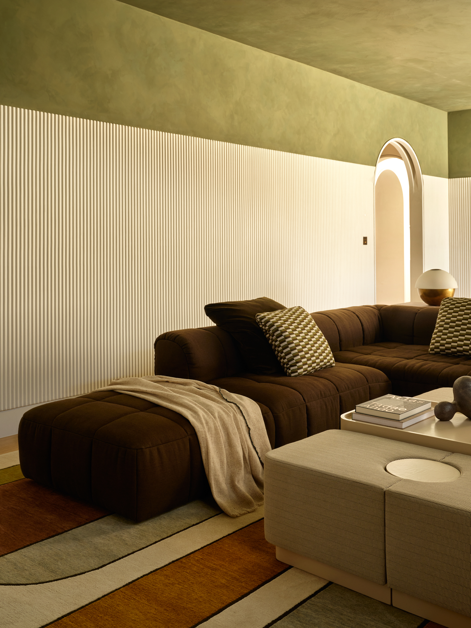 Image of the corner of a living room with walls that are green and white color block. There is a large, dark brown comfy sofa and a colorful rug on the ground.