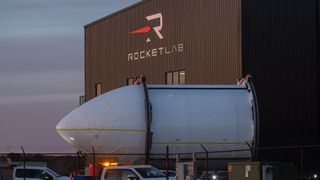 A large, metal bullet-shaped rocket top rests on a trailer in front of a black hangar.