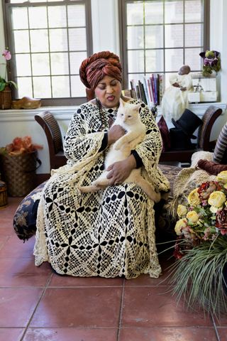 A photo of Marsha Music in an over 60 outfit featuring a crochet gown and red-orange turban and holding her cat.