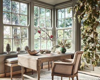 Rustic dining space with large windows overlooking garden, extendable wood dining table, upholstered dining chair and padded bench, with foraged magnolia branch in vase.