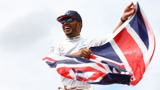 Mercedes driver Lewis Hamilton stands on top of the pitlane fence waving a Union Jack flag to celebrate his win with the British fans ay the F1 British Grand Prix at Silverstone. 