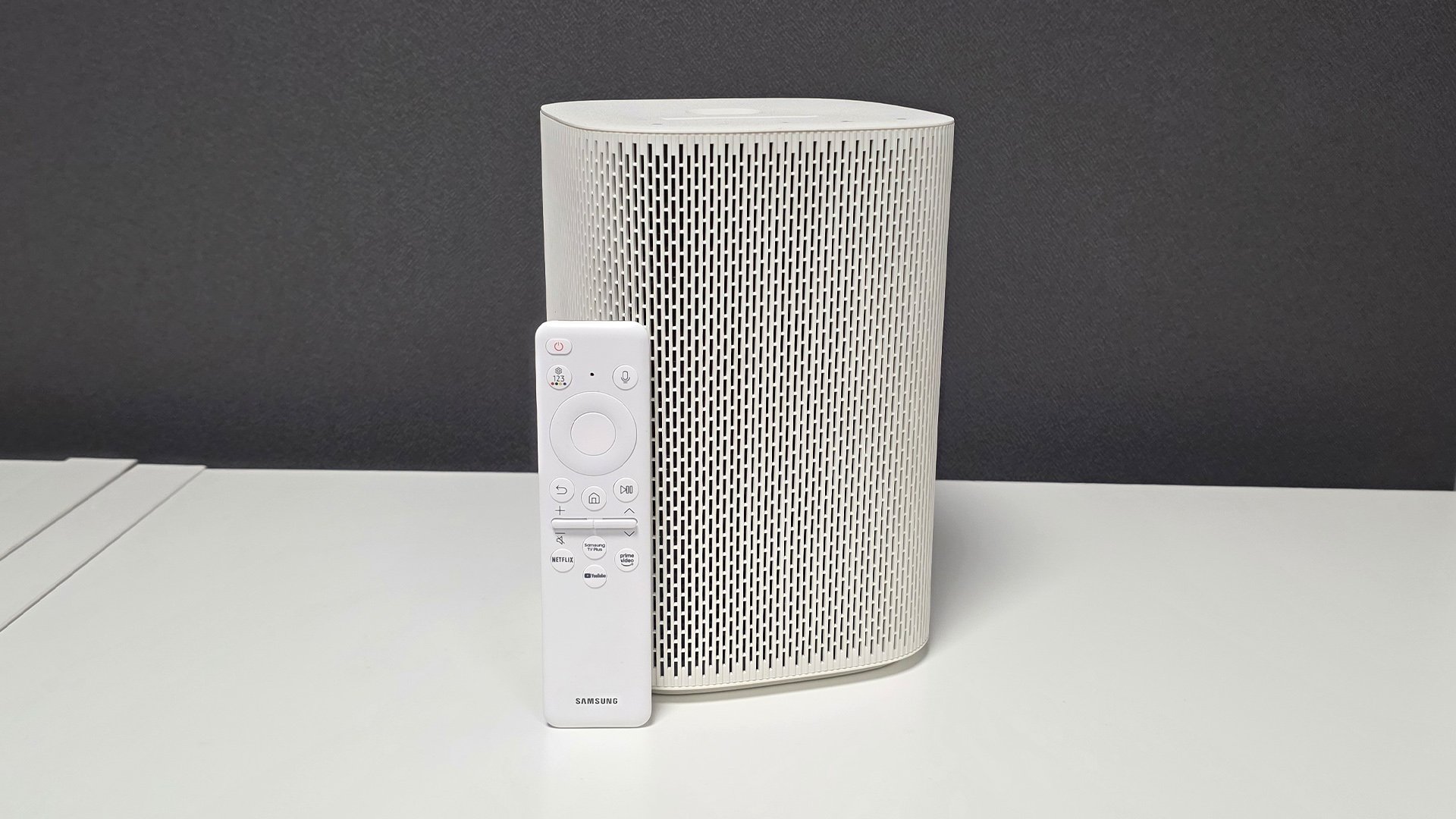 Samsung The Premiere 5 ultra short throw projector on white surface with dark background with remote control propped up against it
