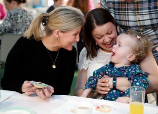 Duchess Sophie wearing a black sweater holding a cookie and smiling at a young girl and her mom at a children's hospice