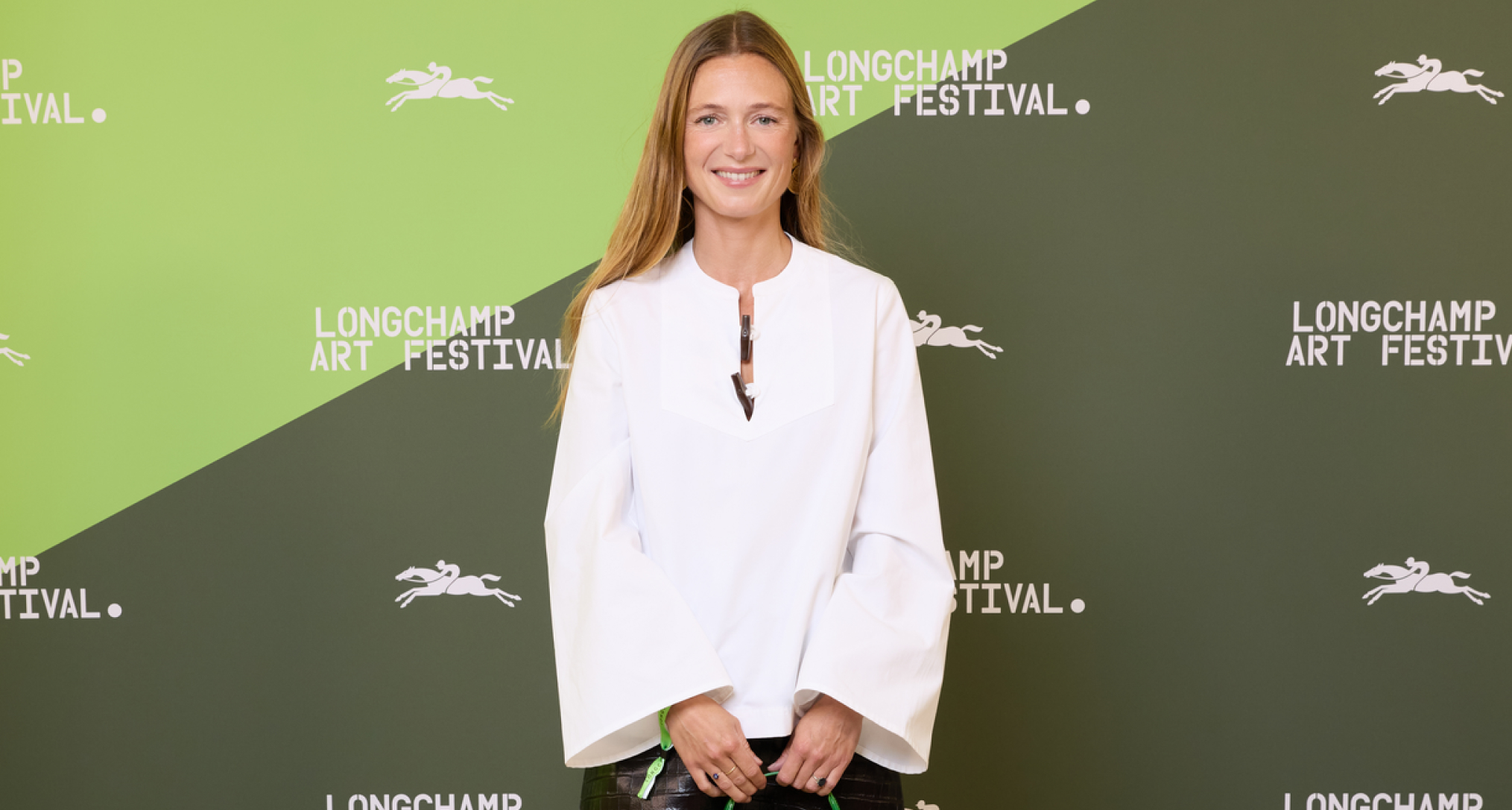 Alizee Thevenet in a white top and leather skirt standing in front of a green Longchamp step and repeat