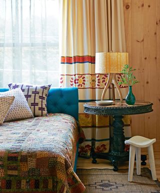bedroom with pine cladding, blue bed with button back headboard, quilt, wooden occasional table and cream patterned curtains