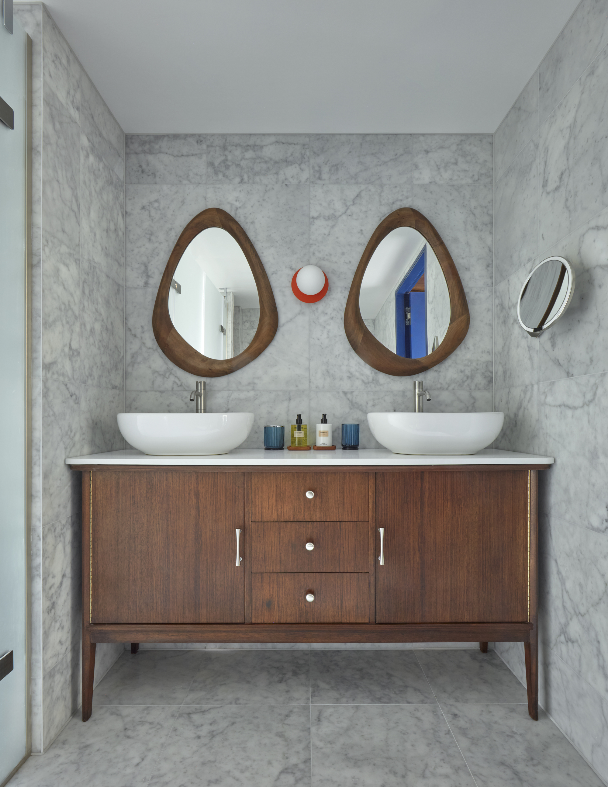 mid century modern inspired bathroom with dark timber freestanding vanity, gray marble tiles on floor and walls, two brown timber accent mirrors, a wall sconce, matching white sinks, silver tapware, a wall mirror and silver hardware