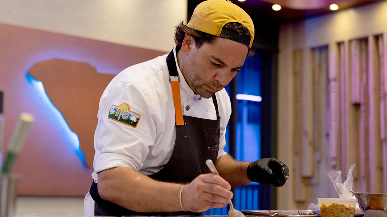 Justin spoons some ambrosia into a cup on Top Chef. He is wearing a yellow hat (and probably shorts).