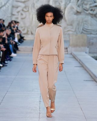 A female model wearing a light brown top and pants and gold sandals walking down a runway.
