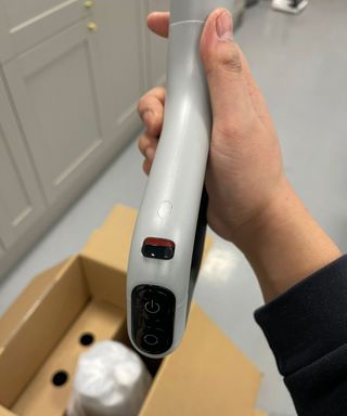 Hand holding the Dreame H14 Pro handle, not attached to the vacuum, showing an on button, mode switch button, and auto distribute switch in front of cardboard box and gray cabinets in Future test center