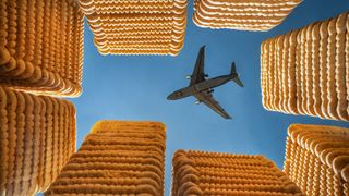 An airplane flies overhead in a blue sky, framed by tall stacks of crackers resembling skyscrapers
