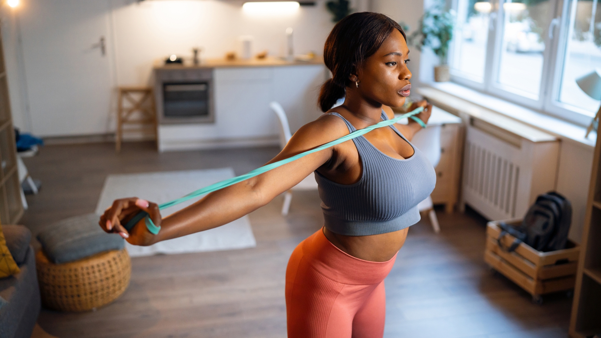 woman holding the ends of a resistance band in each hand and pulling them apart at chest height. she&#039;s in a living room setting looking away from the camera.