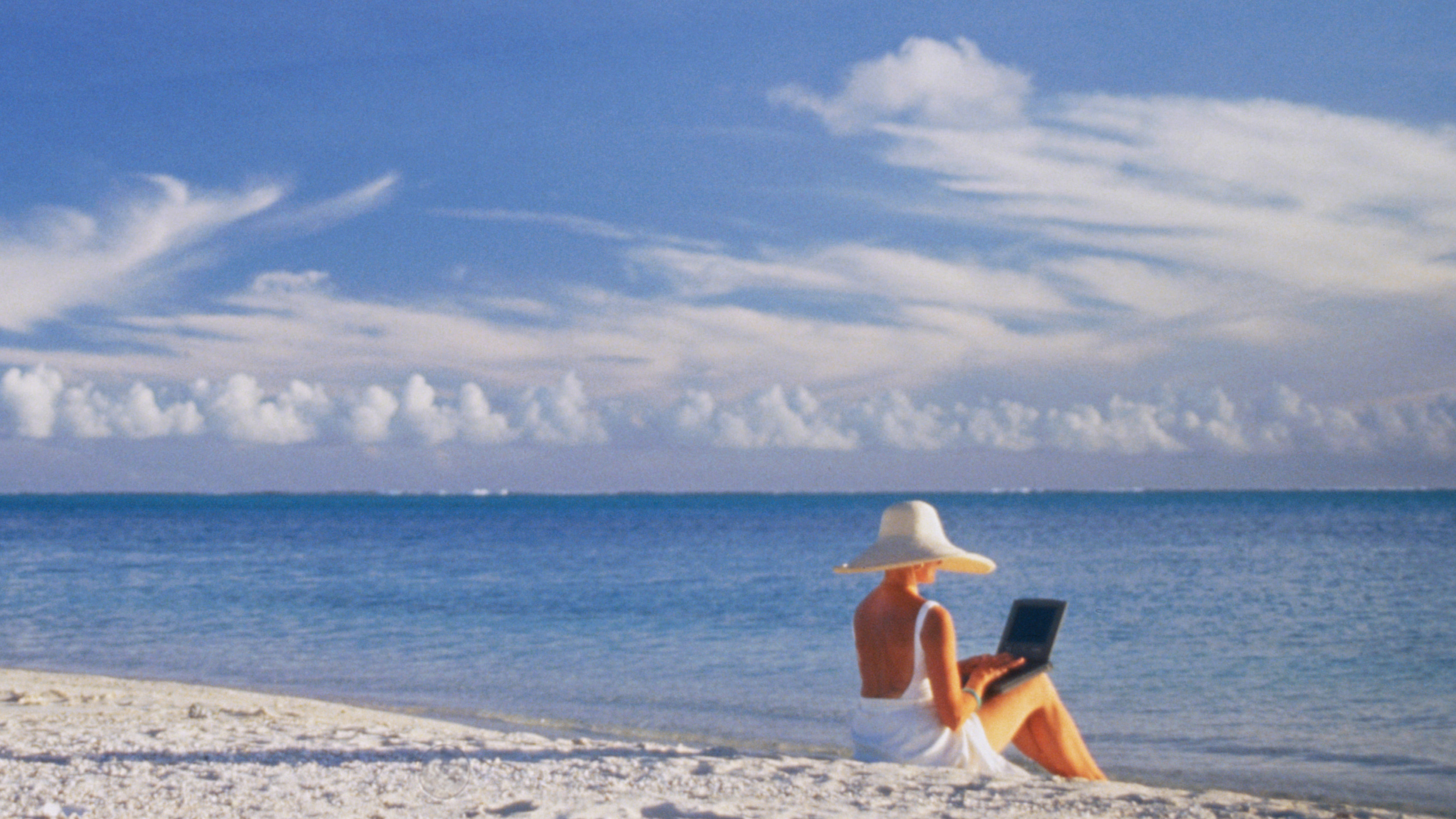 Woman in a white swimsuit, hat and sarong, sitting at the water's edge on a tropical beach and typing on a laptop