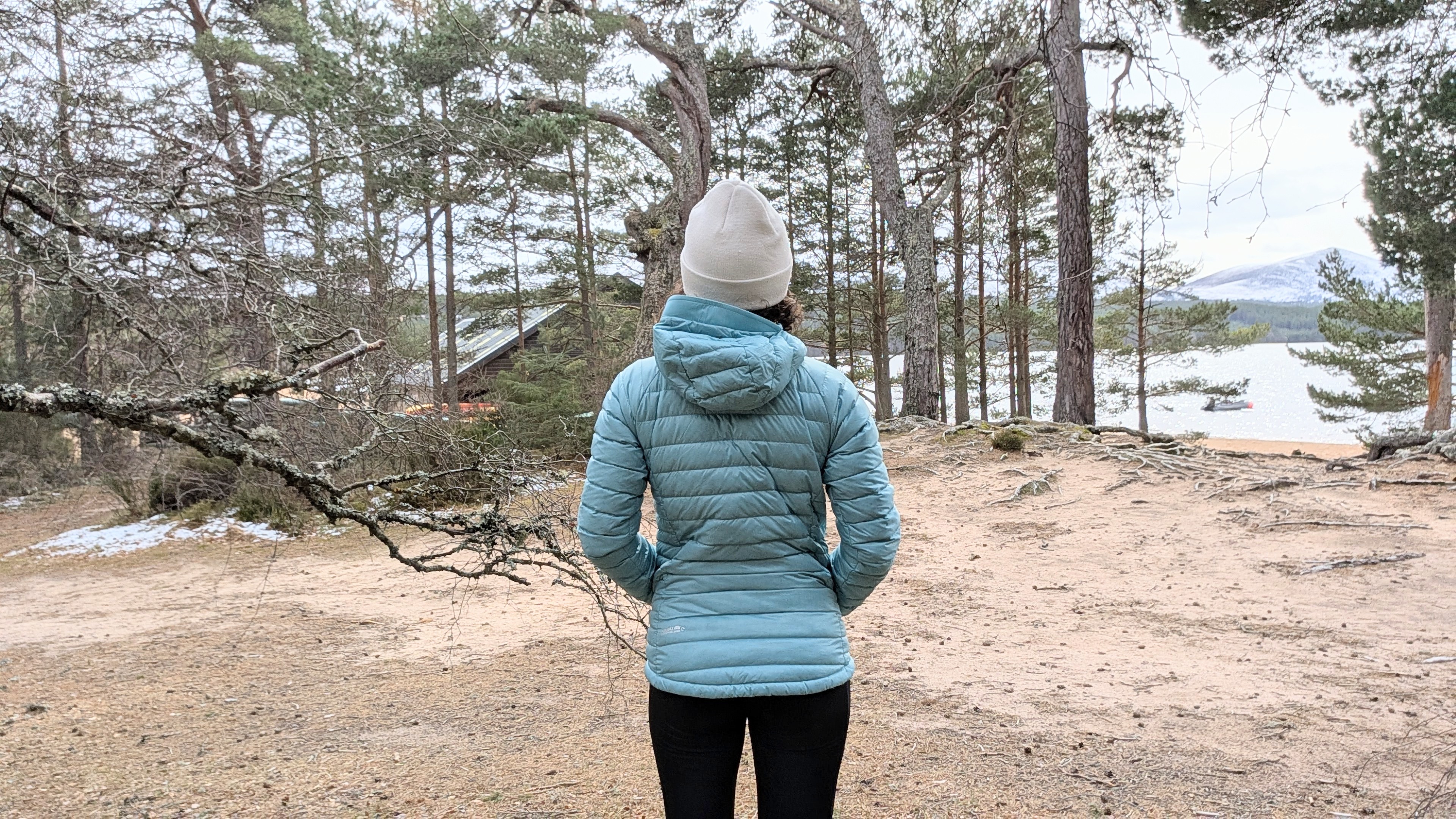 A woman on a beach looking at a lake wearing a bright blue down jacket with ehr hands in her pockets