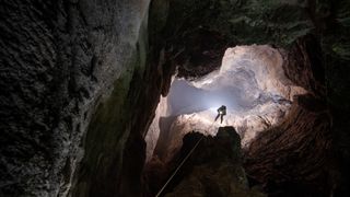 photo looking up at a caver descending into a dark cavern from a bright opening above
