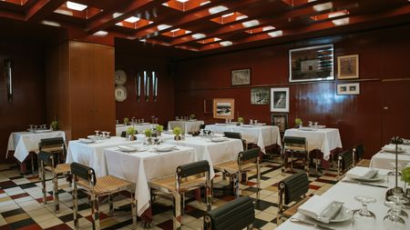 A 1960s-inspired restaurant with coffered, back-lit ceilings, a terracotta-hued palette, checkered floors in red, black, beige, and white, white clothed tables, and rattan, chrome, and leather chairs.